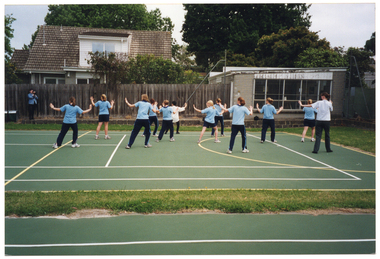 COL photo of a group of students, standing in rows, following a female instructor in some exercises. The students are dressed in their sports uniform, consisting of a blue polo top and either navy blue shorts or track suit pants. The instructor is wearing a cream shirt and black trousers. They are standing on an outdoor tennis courts surrounded by grass. In the background, there is a person wearing a blue shirt and black trousers. They are standing in front of the fence, either Photographing or filming the group. Behind the fence, there is a house and another small building adjoining the property where the fence ends. The Photograph has been taken from behind the group.