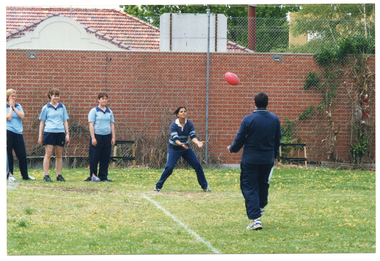 COL photo of a few people standing on a grassy area. They are wearing either pale or dark blue polo shirts and dark blue tracksuit pants. There is a football in mid air between two people, who are facing one another. One of them is standing with her arms positioned in readiness to catch the ball. Beside her, there are three other students standing in a row, awaiting their turn.  They are all standing on a grassy, fenced area, behind which a red brick building is evident.