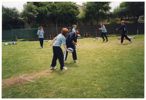 COL photo of a group of people on a fenced grassy area. They are wearing pale or dark blue polo shirts and dark blue tracksuit pants. They are practising handballing and kicking a football with some teachers or instructors. In the background, there are some houses behind a wooden fence.