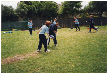 COL photo of a group of people on a fenced grassy area. They are wearing pale or dark blue polo shirts and dark blue tracksuit pants. They are practising handballing and kicking a football with some teachers or instructors. In the background, there are some houses behind a wooden fence.