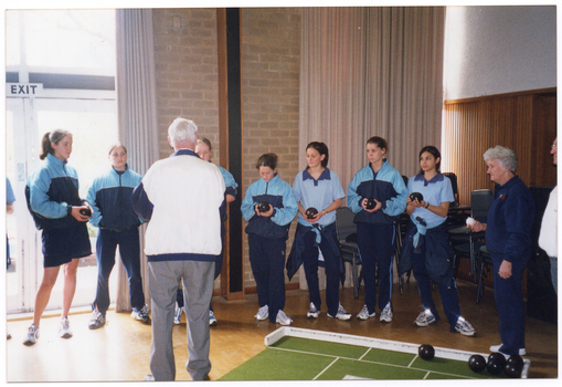 COL photo of a group of teenagers and a few older adults. They are inside a building, where a portable bowling green with a wooden frame at one end has been placed on the parquetry floor. The teenagers are wearing pale blue polo shirts and navy blue tracksuit pants. The adults are wearing navy or white and navy tracksuit jackets over either navy or grey trousers. Most of the teenagers are holding a small black bowling ball in their hands. One older woman is holding a smaller white ball in one hand. There are several black bowling balls on the artificial bowling green on the floor.