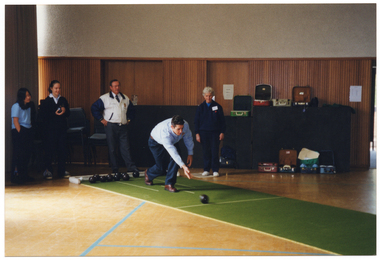 COL photo of a man bowling a small black lawn bowl along a portable artifical bowling green. He is wearing a blue collared shirt and dark trousers. There are four people of varying ages watching him bowl. Two teenagers are  wearing blue polo shirts with navy blue trousers; a middle-aged man is wearing a white shirt and blue tie, with a navy and white tracksuit jacket over navy blue trousers; while an older woman is wearing a navy tracksuit. They are in a school hall or gymnasiun. In the background, some open lawn bowl cases are sitting on top of some black plinths stacked up against a wood-panelled wall.