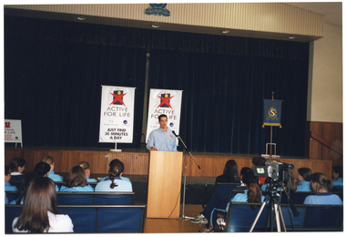 COL photo of a man standing behind a microphone at lectern, on the floor in front of a stage. He is wearing a blue collared shirt. He is addressing a group of teenagers, who are sitting on rows of seats in a hall or gymnasium. The teenagers are all wearing pale blue polo shirts. The man is facing the camera and the teenagers have been Photographed from behind. On the stage behind the man are two freestanding vertical banners, which contain the text, "Active for Life"; an "Active for Life" A-framed sandwich board; and a small Shelford Girls' Grammar School blue and gold banner on a stand.