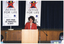 COL photo of a woman standing behind a microphone at lectern, on the floor in front of a stage. She is wearing a red jacket over a blue and white striped t-shirt. Behind her are two freestanding "Active for Life" vertical banners containing the text and logos for Arthritis Victoria and VicHealth. At the bottom of each banner is the text, "JUST FIND 30 MINUTES A DAY".