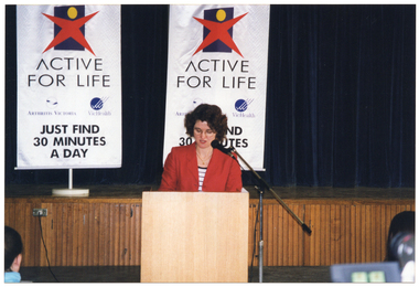 COL photo of a woman standing behind a microphone at lectern, on the floor in front of a stage. She is wearing a red jacket over a blue and white striped t-shirt. Behind her are two freestanding "Active for Life" vertical banners containing the text and logos for Arthritis Victoria and VicHealth. At the bottom of each banner is the text, "JUST FIND 30 MINUTES A DAY".
