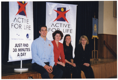 COL photo of four people sitting on the edge of a stage in a hall or gymnasium. Behind them are two freestanding "Active for Life"  vertical banners, with the text and logos for Arthritis Victoria and VicHealth (obscured on one banner). Beneath that is the text "JUST FIND 30 MINUTES A DAY". At the end of the stage, there is a blue and gold Shelford Girls' Grammar School banner on a stand.