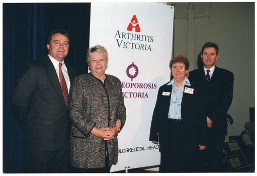 COL photo of four people standing beside a large, vertical, freestanding banner (two on either side). The two men (standing on the outside) are wearing dark suits with a white shirt and tie. One woman (standing centre left) is wearing a mauve top under a black and grey jacket. The other woman (standing centre right) is wearing a pale blue shirt, a black jacket, and black trousers. The banner contains the text and logos for "Arthritis Victoria" and "Osteoporosis Victoria" (partially obscured). At the bottom of the banner, there is some additional text, "MUSCULOSKELETAL HEALTH" (partially obscured). Behind the man standing to the furthest left of the frame, there is a royal blue stage curtain. In the background, behind the two people standing to the right of the frame, there are two two flat decorative columns (colonades) evident on the wall.