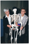 COL photo of two women, standing, posing for a photo with a plastic skeleton on a vertical stand. Both women are wearing white shirts with a jacket - one is wearing a black jacket with dark grey trousers, and the other is wearing a light grey checked jacket with dark trousers. The skeleton is wearing a black hat on its skull.