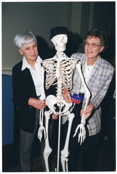 COL photo of two women, standing, posing for a photo with a plastic skeleton on a vertical stand. Both women are wearing white shirts with a jacket - one is wearing a black jacket with dark grey trousers, and the other is wearing a light grey checked jacket with dark trousers. The skeleton is wearing a black hat on its skull.