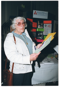 COL photo of a woman standing beside a table. She is wearing an off-white coat over a light grey jumper. She is holding several printed factsheets and brochures in her hands. Behind her, there is a range of printed factsheets on top of the table. At the end of the table, there is a black vertical display board with printed signs and photos on it. At the top of the display board is an "ARTHRITIS VICTORIA" sign. Below that, there are printed signs with text such as, "Telephone Advisory Service", "Self Help Groups", "Self Management Courses", "Library", and "Phone Link". Beneath the "Self Help Groups" sign, there is an information poster for the Melbourne Osteoporosis Support Group. Three photos are evident: a colour photo of three elderly Italian people; a black and white photo of a small group of Vietnamese people; and a colour photo of shelves of books in a library. In the background, there is a potted palm tree (partially obscured) to the left of the frame and an "EXIT" sign above a doorway.