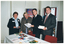 COL photo of four people standing behind and to the side of a rectangular table. One woman is wearing a pale blue shirt, a black jacket, and black trousers. Another woman is wearing a mauve top under a black and grey jacket. The two men are wearing dark suits with a white shirt and tie. On the table, which is covered with a white tablecloth, there is a range of booklets, printed factsheets, and brochures. Three of the people are holding either a booklet, factsheet, or brochure. Behind them are two vertical display boards with a chrome frame. Each display board has some signs or posters on it.