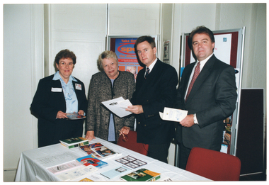 COL photo of four people standing behind and to the side of a rectangular table. One woman is wearing a pale blue shirt, a black jacket, and black trousers. Another woman is wearing a mauve top under a black and grey jacket. The two men are wearing dark suits with a white shirt and tie. On the table, which is covered with a white tablecloth, there is a range of booklets, printed factsheets, and brochures. Three of the people are holding either a booklet, factsheet, or brochure. Behind them are two vertical display boards with a chrome frame. Each display board has some signs or posters on it.