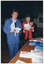 COL photo of two women standing in front of a table. One woman is wearing a blue leisure suit with a pale blue top. The other woman is wearing a red jacket over a white top and black trousers. They are each holding an open book. On top of the table, there are various books on display. In the background, there is a medium sized vertical banner on a stand. It contains the text and logo for the City of Glen Eira.