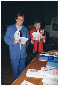 COL photo of two women standing in front of a table. One woman is wearing a blue leisure suit with a pale blue top. The other woman is wearing a red jacket over a white top and black trousers. They are each holding an open book. On top of the table, there are various books on display. In the background, there is a medium sized vertical banner on a stand. It contains the text and logo for the City of Glen Eira.