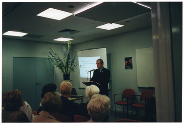 COL photo of a man speaking into a microphone at a lectern. Behind him is a projector screen displaying a slide image titled, "Osteoporosis 2002 FAQs - Frequently Asked Questions". In front of him is a small table with a laptop computer. To the side, there is a large bouquet of flowers in a vase on a stand. On the wall, there is a poster with the image of two hands holding a wishbone, with the text "WORLD OSTEOPOROSIS DAY" below. In the foreground, some members of the seated audience have been Photographed from behind.
