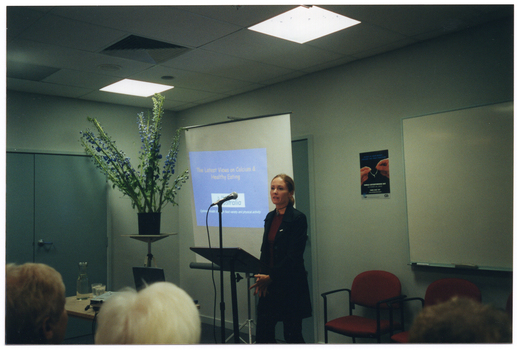 COL photo of a woman speaking into a microphone at a lectern. Behind her is a projector screen displaying a slide image titled, "The Latest Views on Calcium & Healthy Eating". To the side, there is a large bouquet of flowers in a vase on a stand. On the wall, there is a poster with the image of two hands holding a wishbone, with the text "WORLD OSTEOPOROSIS DAY" below.