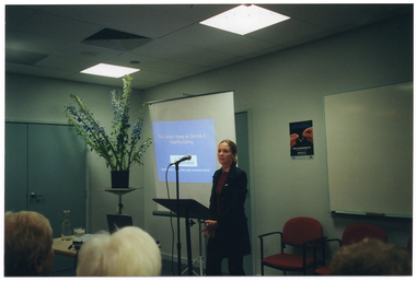 COL photo of a woman speaking into a microphone at a lectern. Behind her is a projector screen displaying a slide image titled, "The Latest Views on Calcium & Healthy Eating". To the side, there is a large bouquet of flowers in a vase on a stand. On the wall, there is a poster with the image of two hands holding a wishbone, with the text "WORLD OSTEOPOROSIS DAY" below.