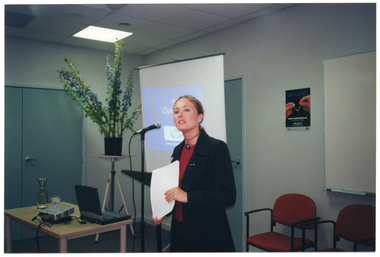 COL photo of a woman speaking into a microphone beside a lectern. Behind her is a projector screen displaying a slide image titled, "Any Questions?" (partially obscured). Beside her is a small table with a laptop computer, a slide projector, a glass carafe and two glasses of water. Behind the table, there is a large bouquet of flowers in a vase on a stand. On the wall, there is a poster with the image of two hands holding a wishbone, with the text "WORLD OSTEOPOROSIS DAY" below.