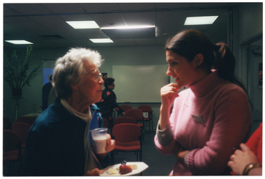 COL medium close up, slightly blurred photo of three women speaking to one another in a room, (one woman is almost completely obscured). One of the women is holding a plastic cup of milky tea. There are two other people in the background. There are chairs placed in rows facing a large whiteboard affixed to the wall, and a freestanding slide projector screen to one side of the whiteboard. Also in the background, to the left of the frame, there is a large bouquet of flowers in a vase on a stand.