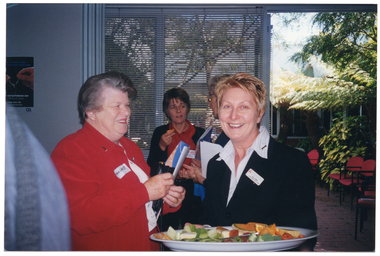 COL medium close up photo of three women in a room. Two of the women are standing in the foreground, close to one another. One of them is carrying a printed booklet and a wallet or small bag in her hands. The other woman is carrying a large fruit platter. In the background, there is a third woman standing further behind, in front of a large window with thin venetian blinds. She is holding an orange wedge in one hand. Beside the window, there is an open doorway leading to a courtyard with ferns and other plants. Some red/burgundy chairs in the courtyard have been placed in the courtyard. On the wall near the window, there is a poster with the image of two hands holding a wishbone, with the text "WORLD OSTEOPOROSIS DAY". In the foreground, to the left of the frame, the back of a person (mostly obscured) is walking away.