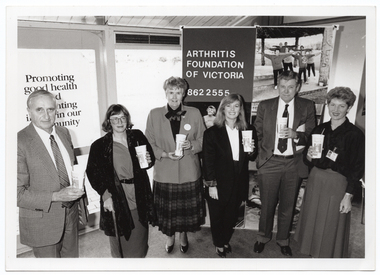 B&W photo of a small group of people standing in a semi-circle in front of a large window and a freestanding vertical display board. They are each holding a disposable PhysiCal branded milk-shake cup with a straw. There is a large poster on one of the windows, which contains the text (partially obscured), "Promoting good health and preventing illness in our community". On one panel of the display board, there is the text, "Arthritis Foundation of Victoria 862 2555", and some large posters of people doing various types of exercises, including water exercise.