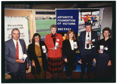 COL photo of a small group of people standing in a semi-circle in front of a large window and a freestanding vertical display board. They are each holding a disposable PhysiCal branded milk-shake cup with a straw. There is a large poster on one of the windows, which contains the text (partially obscured), "Promoting good health and preventing illness in our community". On one panel of the display board, there is the text, "Arthritis Foundation of Victoria 862 2555", and some large posters of people doing various types of exercises, including water exercise. Through the window, there is a view of a grassy sports oval, with a row of houses and some trees in the distance.