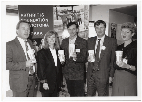 B&W photo of a small group of people standing in a semi-circle in front of a large freestanding vertical display board. They are each holding a disposable PhysiCal branded milk-shake cup with a straw. On the left panel of the display board, there is the text, "Arthritis Foundation of Victoria 862 2555". On the right panel, there is a photo of a group of people exercising.