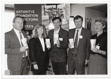 B&W photo of a small group of people standing in a semi-circle in front of a large freestanding vertical display board. They are each holding a disposable PhysiCal branded milk-shake cup with a straw. On the left panel of the display board, there is the text, "Arthritis Foundation of Victoria 862 2555". On the right panel, there is a photo of a group of people exercising.