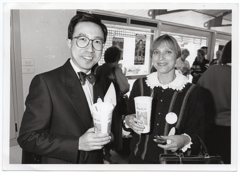 B&W medium close up photo of a man and a woman posing for a photo. They are each holding a PhysiCal branded disposable milkshake cup with a straw. The woman is also holding an hors d'oeuvre in her other hand. In the background, there are several other people standing and chatting. One woman is drinking from a cup. There are some posters on the windows, beyond which some wooden lattice panels are evident around the outside of the windows.