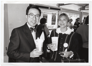 B&W medium close up photo of a man and a woman posing for a photo. They are each holding a PhysiCal branded disposable milkshake cup with a straw. The woman is also holding an hors d'oeuvre in her other hand. In the background, there are several other people standing and chatting. One woman is drinking from a cup. There are some posters on the windows, beyond which some wooden lattice panels are evident around the outside of the windows.