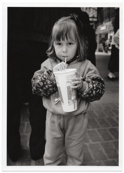 B&W medium close up photo of a young girl drinking through a straw from a PhysiCal branded disposable milkshake cup. She is standing outdoors on a paved brick floor. There is a crowd of people behind her. Through a gap in the crowd, some shops are evident in the distance.