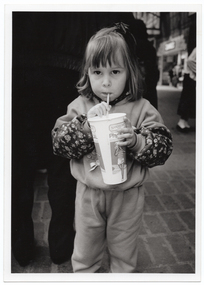 B&W medium close up photo of a young girl drinking through a straw from a PhysiCal branded disposable milkshake cup. She is standing outdoors on a paved brick floor. There is a crowd of people behind her. Through a gap in the crowd, some shops are evident in the distance.