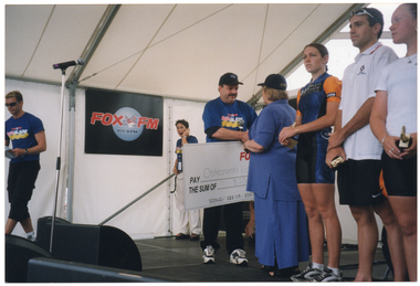 COL photo of a woman accepting a large novelty cheque from a man. He is wearing a black cap with the Paul's ice cream text and logo, and a blue t-shirt containing the "Fox FM" and PhysiCal Blade text and logos. She is wearing a matching blue dress and shirt. The cheque is made out to Osteoporosis Victoria for $2000. They are standing on a stage, under the cover of a large marquee. Two people appearing in the background are also wearing the same sponsor branded t-shirt - one is speaking into an ear-piece and the other is walking off the stage. Beside the woman, there are three people standing in a row, each holding a small trophy. One is wearing a lycra top and shorts, while the other two are wearing polo shirts and shorts.