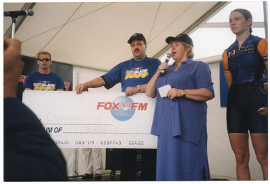 COL photo of a woman speaking into a microphone. Behind her, a man is holding up a large novelty cheque. He is wearing a black cap with the Paul's ice cream text and logo, and a blue t-shirt containing the "Fox FM" and PhysiCal Blade text and logos. The cheque is made out to Osteoporosis Victoria for $2000. They are on a stage, under the cover of a large marquee. Standing beside her is a sports person dressed in a lycra top and shorts. Standing in the background is another man wearing the same sponsor branded t-shirt and another person (obscurred by the large novelty cheque). In the foreground, to the left of the frame, another person's hand and arm can be seen, positioned as if taking a photo of the people onstage (although the camera itself is not visible).