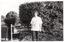 B&W photo of a woman standing beside a large wooden sign. The sign is situated in front of some trees and shrubs, beside a road or driveway. Carved into the wooden sign is the text "A.F.V. ARTHRITIS CENTRE" (partially obscured). The woman is wearing a t-shirt with a "Joint Action" logo printed on the front. In the distance, there is a car to the left of the frame.