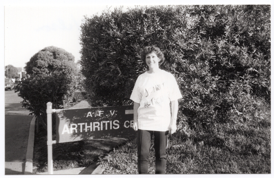 B&W photo of a woman standing beside a large wooden sign. The sign is situated in front of some trees and shrubs, beside a road or driveway. Carved into the wooden sign is the text "A.F.V. ARTHRITIS CENTRE" (partially obscured). The woman is wearing a t-shirt with a "Joint Action" logo printed on the front. In the distance, there is a car to the left of the frame.