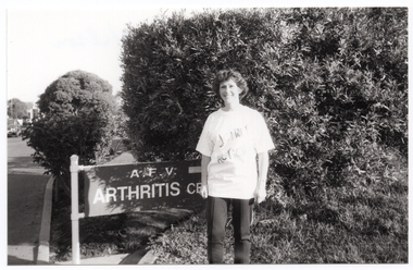 B&W photo of a woman standing beside a large wooden sign. The sign is situated in front of some trees and shrubs, beside a road or driveway. Carved into the wooden sign is the text "A.F.V. ARTHRITIS CENTRE" (partially obscured). The woman is wearing a t-shirt with a "Joint Action" logo printed on the front. In the distance, there is a car to the left of the frame.