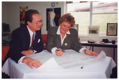 COL photo of two people sitting at a small table. They are looking down at a draft of some building plans. In the background, there is a computer, some posters on the wall, and some framed prints and certificates on the window sill. Through the window, there is a view of another building close by, situated at a higher level on a grassy hill.