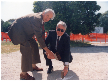 COL photo of two men posing for a photo with a mallet above a large wooden peg that has been hammered into the ground. They are both holding the mallet above the peg, while one of the men also has his other hand on the peg. In the background, there is a bright orange plastic barrier mesh safety fence, which has been erected along the street boundary of the property. Across the road, there is a sign with the text, "CAULFIELD GENERAL MEDICAL CENTRE" (partially obscured).