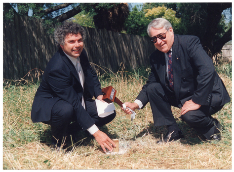 COL photo of two men posing for a photo on a grassy area. One of the men is holding a wooden mallet aloft above a large wooden peg that has been hammered into the ground. The other man has his hand on the peg. In the background, there is a wooden fence, behind which there is a large tree.