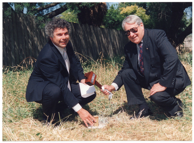 COL photo of two men posing for a photo on a grassy area. One of the men is holding a wooden mallet aloft above a large wooden peg that has been hammered into the ground. The other man has his hand on the peg. In the background, there is a wooden fence, behind which there is a large tree.