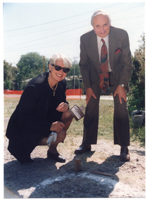 COL photo of two people posing for a photo on a grassy area. The woman is holding a wooden mallet above a large wooden peg that has been hammered into the ground. A man is standing next to her, bending forward with his hands on his knees. In the background, a bright orange plastic barrier mesh safety fence, which has been erected along the street boundary of the property, is evident. There is also a building evident across the road, behind some shrubs.