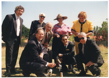 COL photo of a group of people, some holding wooden mallets, posing for a photo around a large wooden peg which has been hammered into the ground. One man is holding a wooden mallet above the peg.