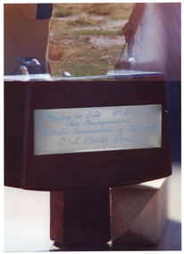 COL close up photo of a wooden mallet, with a wooden peg lying on its side beneath it. There is a small metal plaque on the mallet, engraved with the text, " "Pegging the site" 8th December '93 New Headquarters Arthritis Foundation of Victoria H & L Hecht Trust".