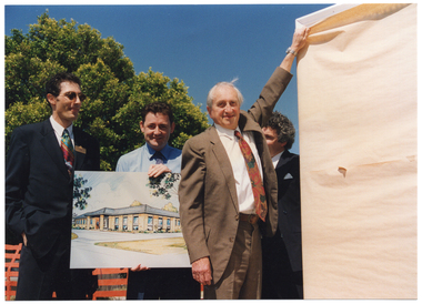 COL photo of four men standing beside a tall rectangular sign covered in brown paper. One of the men is holding a colour illustration of a building. Another one of the men is reaching up to the top left corner of the sign to pull the brown paper covering down from the sign. One of the men is standing behind him (partially obscured).