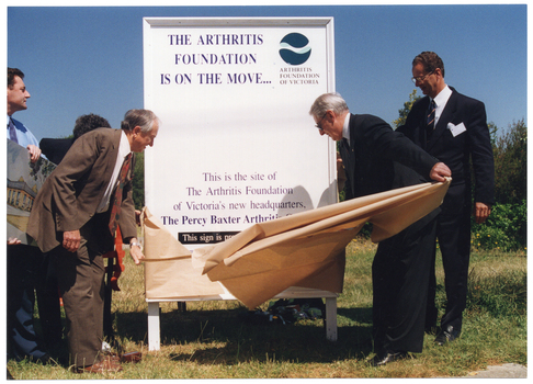 COL photo of two men pulling a brown paper covering off a large advertising billboard that has been affixed to the ground. There are three other men beside them (one partially obscured). One of them is holding a colour illustration of a building. The text on the billboard says, "THE ARTHRITIS FOUNDATION IS ON THE MOVE... This is the site of the Arthritis Foundation of Victoria's new headquarters, The Percy Baxter Arthritis Centre" (partially obscured). This sign is a pr..." (partially obscured). The billboard has been affixed to the ground on a vacant block of land.
