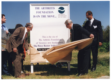COL photo of two men pulling a brown paper covering off a large advertising billboard that has been affixed to the ground. There are three other men beside them (one partially obscured). One of them is holding a colour illustration of a building. The text on the billboard says, "THE ARTHRITIS FOUNDATION IS ON THE MOVE... This is the site of the Arthritis Foundation of Victoria's new headquarters, The Percy Baxter Arthritis Centre" (partially obscured). This sign is a pr..." (partially obscured). The billboard has been affixed to the ground on a vacant block of land.