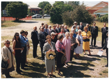 COL photo of a group of people gathered outdoors on a vacant block of land. They are all looking in the same direction. To the right of the frame, there is a man facing the group. He is holding a piece of paper. In the background, there is a bright orange plastic barrier mesh safety fence, which has been erected along the street boundary of the property, and several shrubs. There is a taxi travelling along the road, and some buildings across the road.