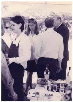 B&W medium close up photo of a few people gathered under a marquee, outdoors. Three people are in a small group, chatting. A waitress, wearing a dark coloured apron, is holding a tray of refreshments. She has her back to the small group. Another two people are evident on the left side edge of the frame. On a table in the foreground are bottles of wine, flute champagne glasses, and a bowl of strawberries.