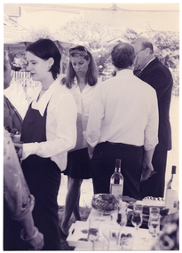 B&W medium close up photo of a few people gathered under a marquee, outdoors. Three people are in a small group, chatting. A waitress, wearing a dark coloured apron, is holding a tray of refreshments. She has her back to the small group. Another two people are evident on the left side edge of the frame. On a table in the foreground are bottles of wine, flute champagne glasses, and a bowl of strawberries.